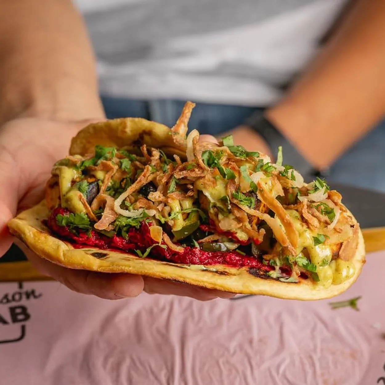 Hand holding a loaded flatbread kebab on a Maison Bab tray