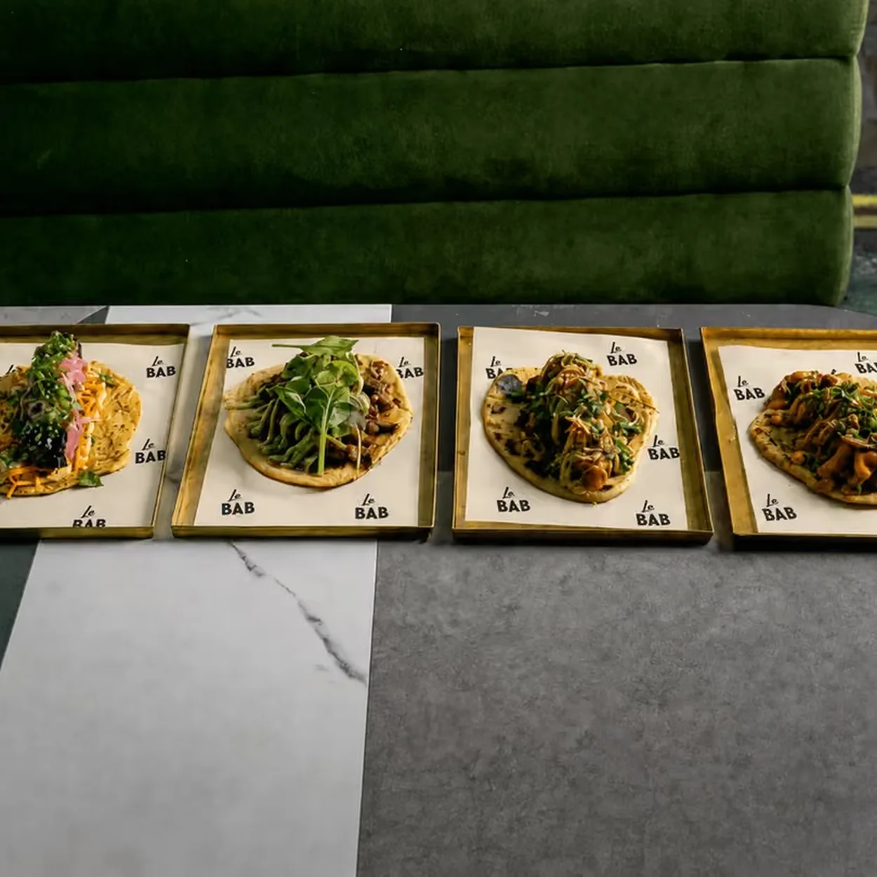 Four kebabs on metal trays lined up on a table in front of a green banquette