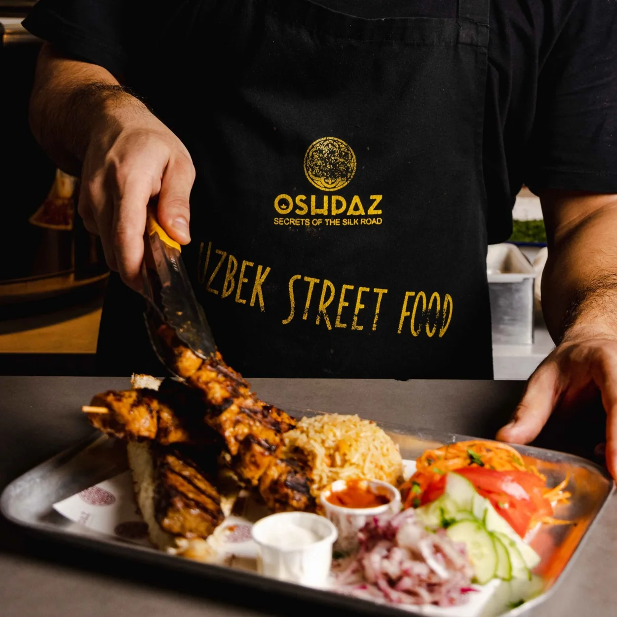 OshPaz member of staff in a branded Uzbek Street Food apron serving a tray of grilled skewers, rice and salad