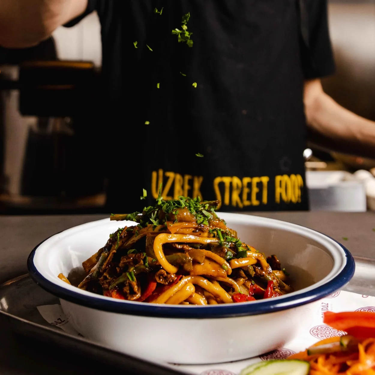 OshPaz bowl of Uzbek noodles with peppers and herbs on the counter with the kitchen behind
