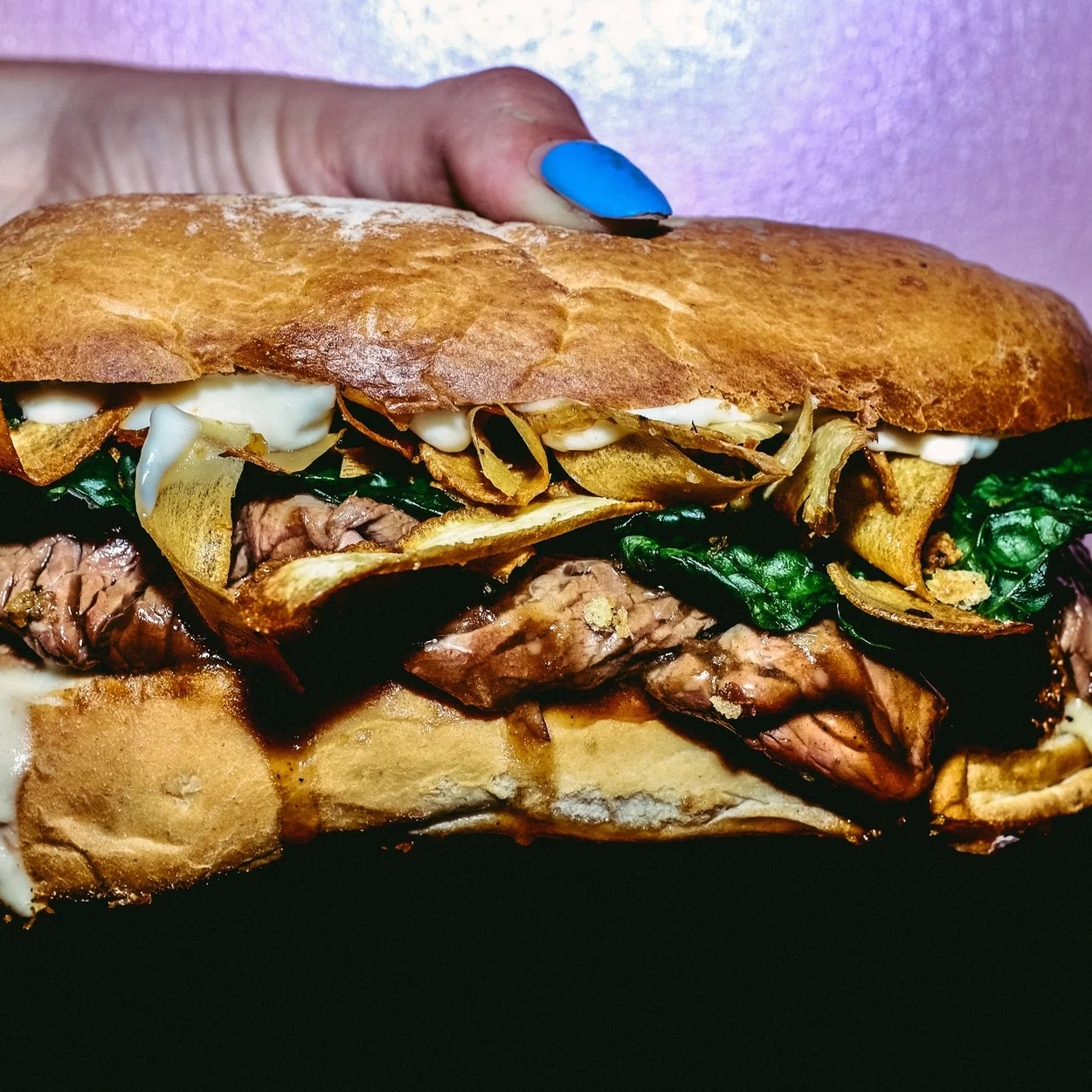 STAKEHAUS steak sandwich layered with melted cheese and greens held against a purple background